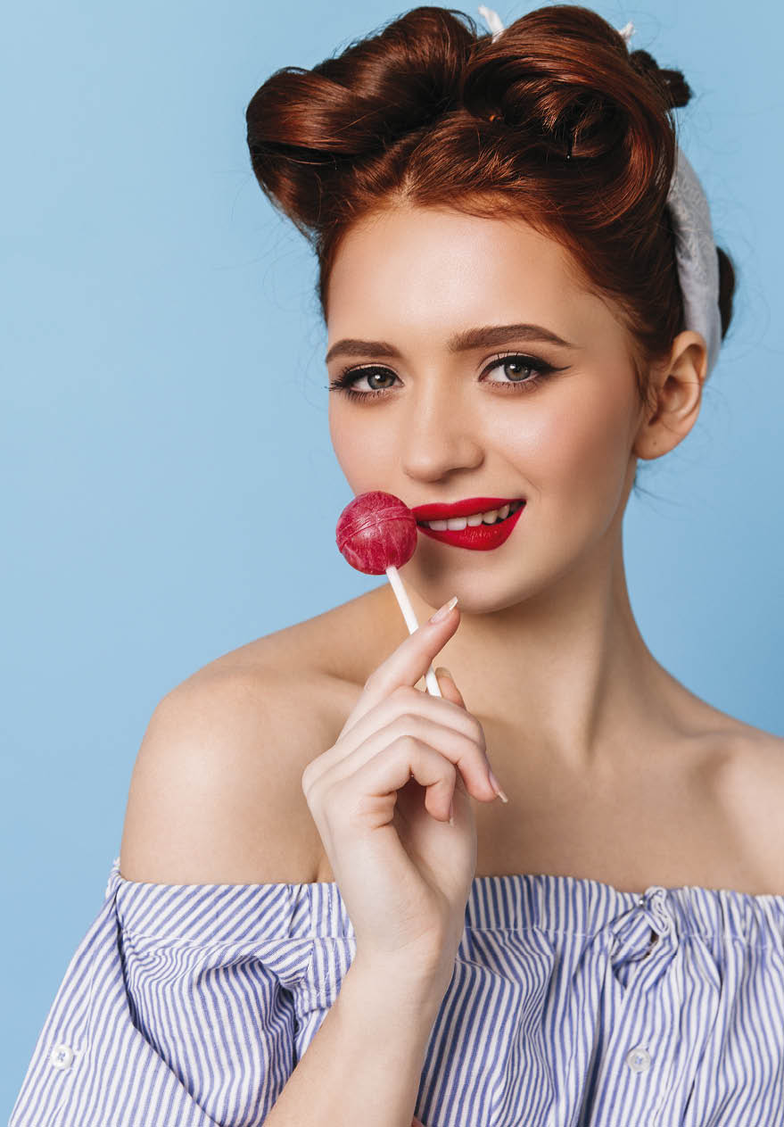 Playful ginger woman licking hard candy. Studio shot of glamorous pinup girl with lollipop isolated on blue background.